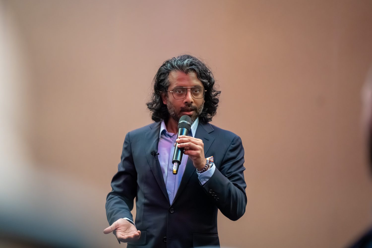 Speaker mid-talk with an open, palm-up gesture to the audience