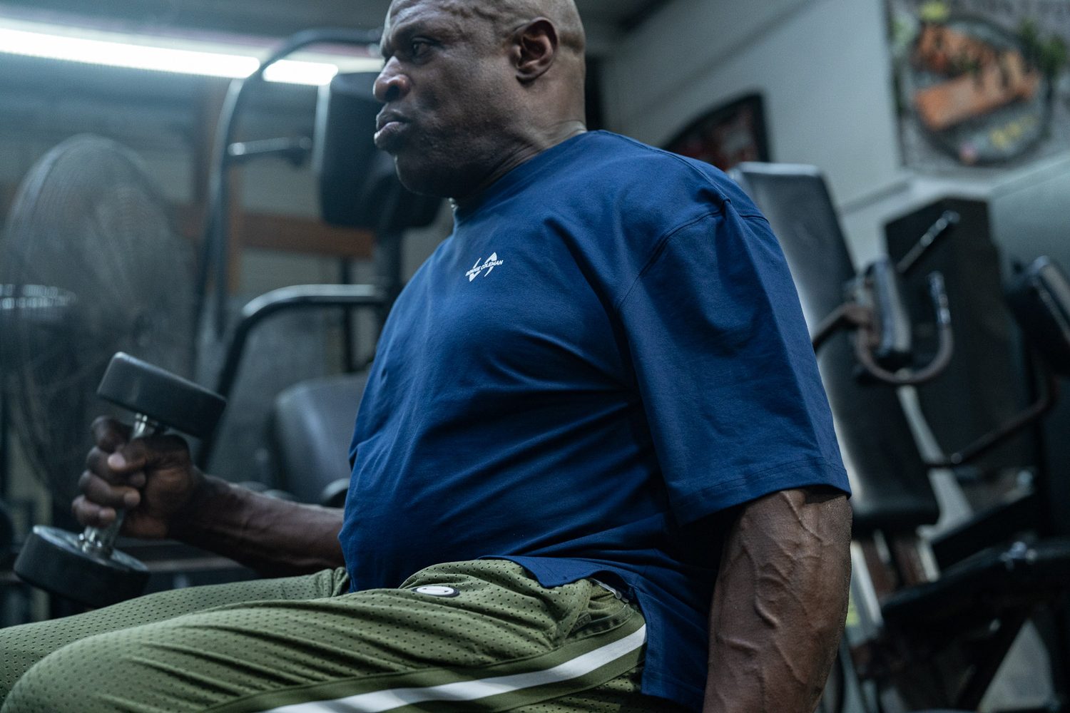 Ronnie Coleman seated with a dumbbell in side profile under gym lights